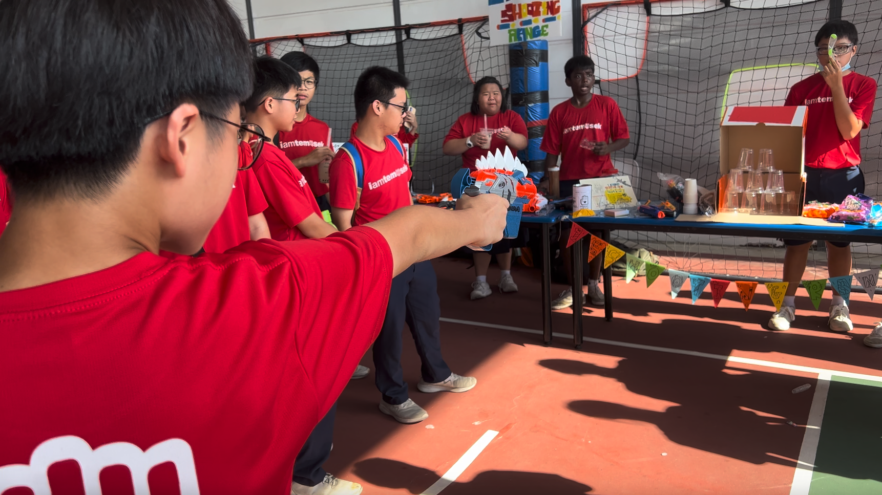 A person in a red shirt aims a toy foam blaster at a shooting range game booth at an outdoor event.
